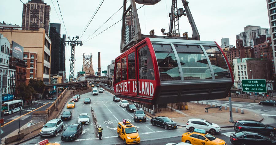 The Roosevelt Island Tramway, painted a distinct red with big white lettering on the side, travels across cables over a busy NYC street.