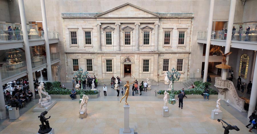 People explore The American Wing of The Met in New York City, where several marble and metal statues are illuminated by natural light through the glass ceiling.