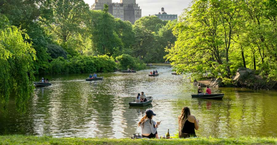 Two women picnic on the grassy bank of The Lake in Central Park, watching rowboats on the water and enjoying spring in NYC.