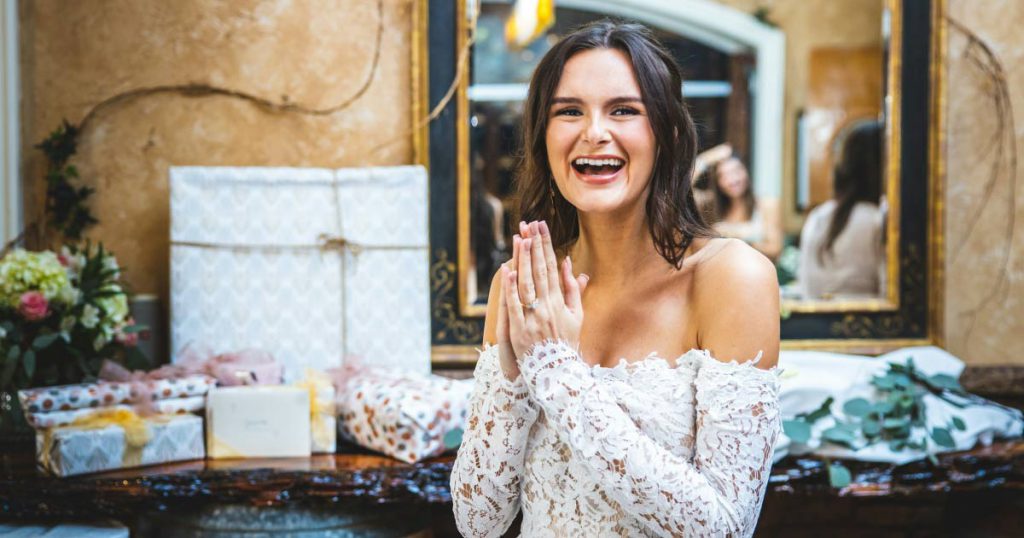 A woman in a white lace dress is holding her hands together and smiling widely beside a table of bridal shower gifts.