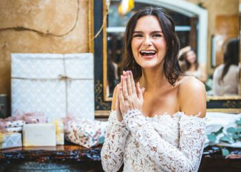 A woman in a white lace dress is holding her hands together and smiling widely beside a table of bridal shower gifts.