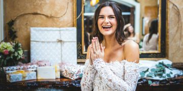 A woman in a white lace dress is holding her hands together and smiling widely beside a table of bridal shower gifts.
