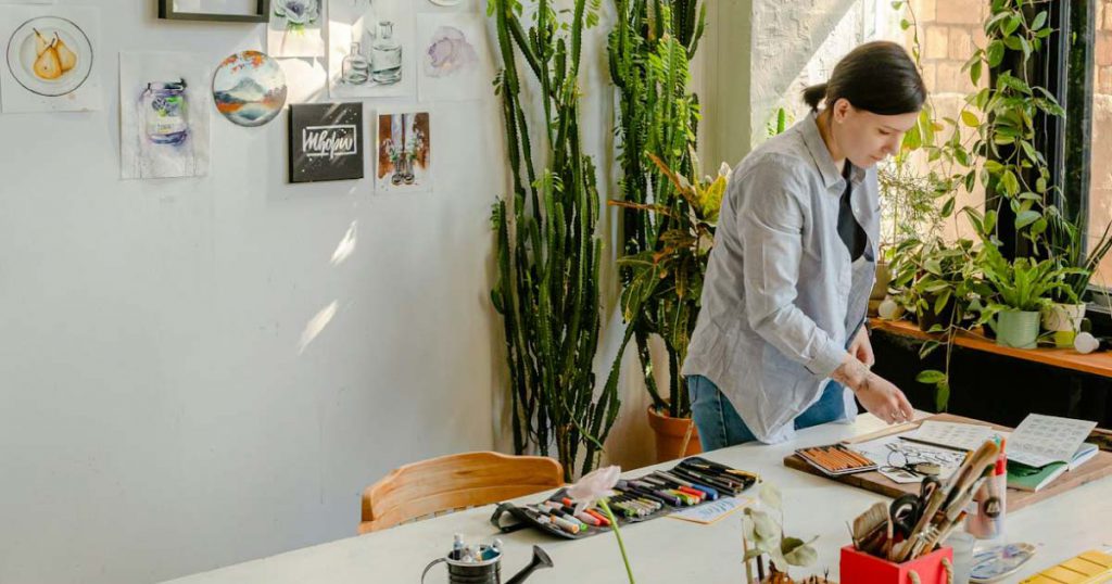 An artist is looking at papers on the workbench in her home art studio, surrounded by houseplants, drawings, and paintings.