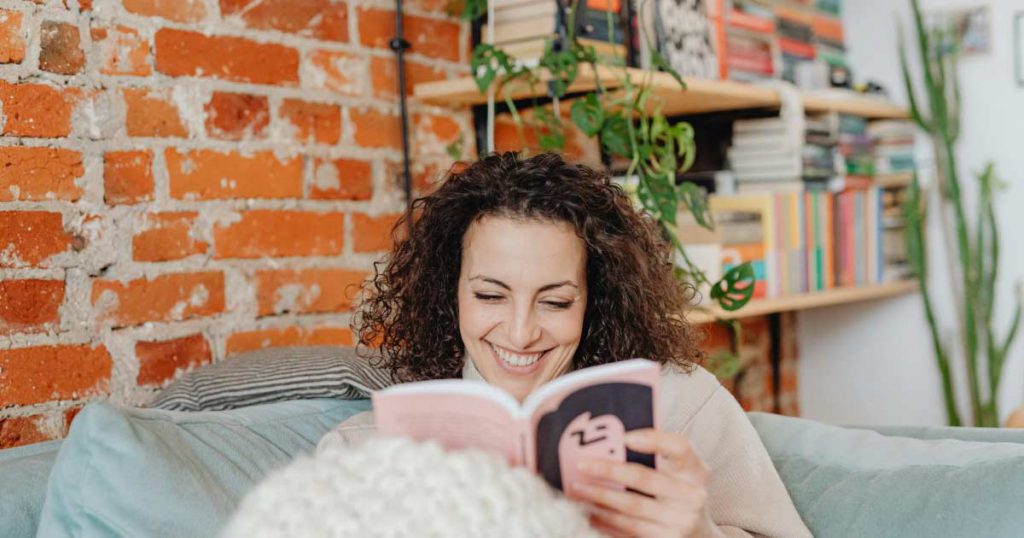 A woman is sitting on her couch under a fuzzy blanket, reading a book in a cozy reading nook.