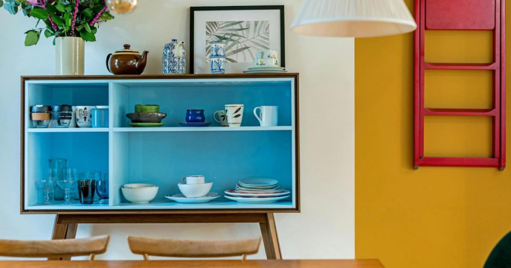 View over a kitchen table of a blue dish cupboard set against a white wall with a stark goldenrod accent, showcasing a couple of ways to create a colorful home.