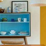 View over a kitchen table of a blue dish cupboard set against a white wall with a stark goldenrod accent, showcasing a couple of ways to create a colorful home.