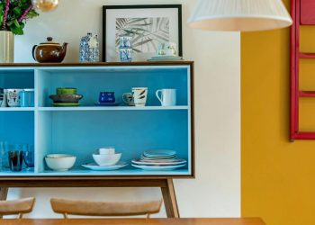 View over a kitchen table of a blue dish cupboard set against a white wall with a stark goldenrod accent, showcasing a couple of ways to create a colorful home.