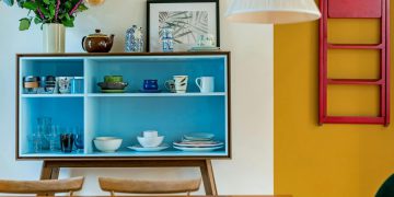 View over a kitchen table of a blue dish cupboard set against a white wall with a stark goldenrod accent, showcasing a couple of ways to create a colorful home.