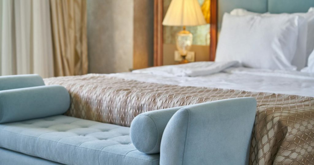 A bedroom with cool blue accents in the form of an upholstered bench at the foot of the bed and a padded headboard.