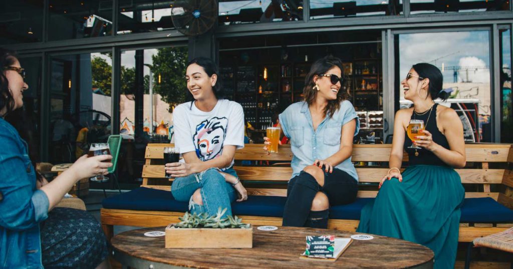 Four women are laughing together, enjoying sunny weather and craft beers at a local brewpub in Fort Lauderdale.