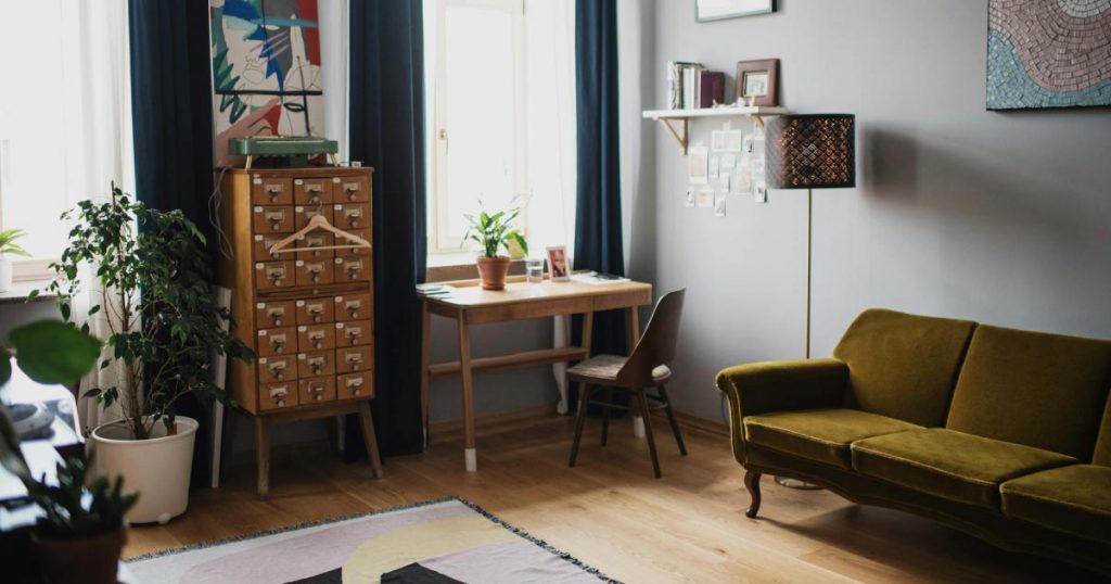 A cozy living room, featuring a simple desk, house plants, an abstract rug, and a mid-century modern sofa in an earthy green tone.