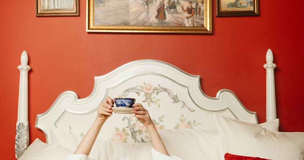 View of a room decorated with persimmon-colored walls and gold-framed artwork, featuring a person lying in bed holding up an elegant teacup against an ornate white headboard.