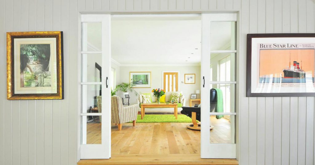 French doors open to a bright living room in a colorful home, with a wasabi-colored rug, natural wood floors, and several pieces of art hanging on the walls.