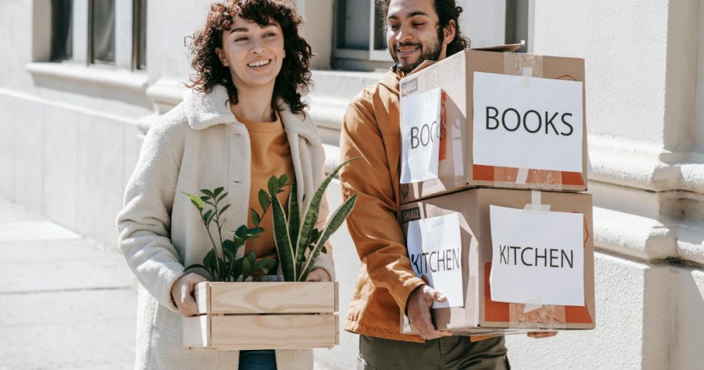 A happy couple is walking to their car with moving boxes labeled “KITCHEN” and “BOOKS” and a small wooden crate of houseplants.