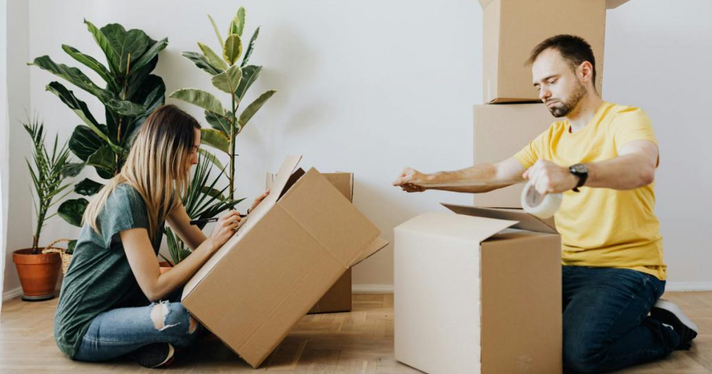 A man and woman are sitting on the floor beside a few tall houseplants, assembling cardboard boxes for moving plants.