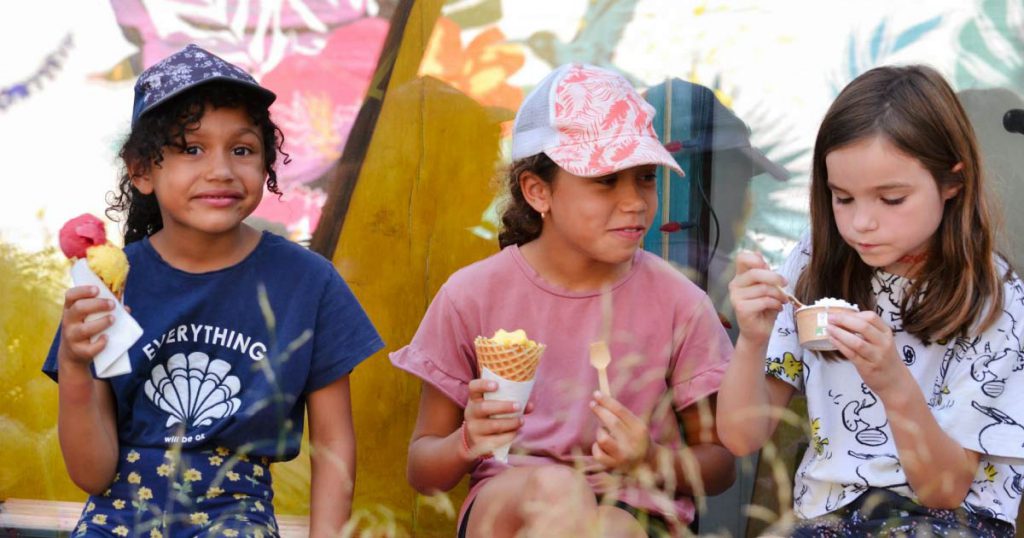Three young girls are sitting in front of a colorful muraled wall while enjoying ice cream in Fort Lauderdale on a sunny day.