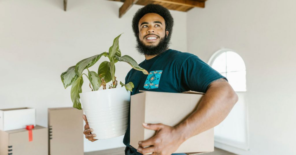 A happy man who knows how to move with plants is holding a houseplant in one arm and a moving box in the other.