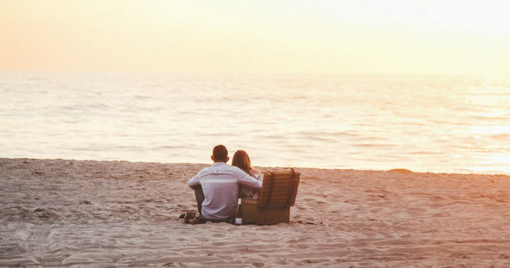 A couple is sitting in the sand beside a picnic basket while watching the sunset at a beach in Fort Lauderdale, Florida.