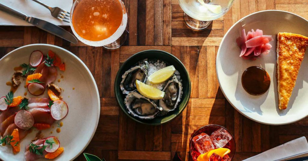 View from above of a table set with high-end food and drink, including raw oysters, sashimi, a slice of cake, and three cocktails.