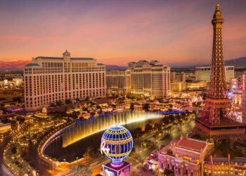 Aerial view of the Las Vegas Strip skyline at sunset featuring the illuminated Bellagio fountains, Eiffel Tower replica, and mountains in the distance.