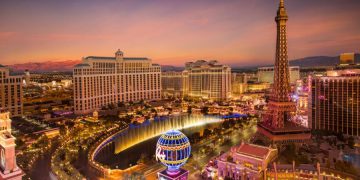 Aerial view of the Las Vegas Strip skyline at sunset featuring the illuminated Bellagio fountains, Eiffel Tower replica, and mountains in the distance.