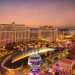 Aerial view of the Las Vegas Strip skyline at sunset featuring the illuminated Bellagio fountains, Eiffel Tower replica, and mountains in the distance.