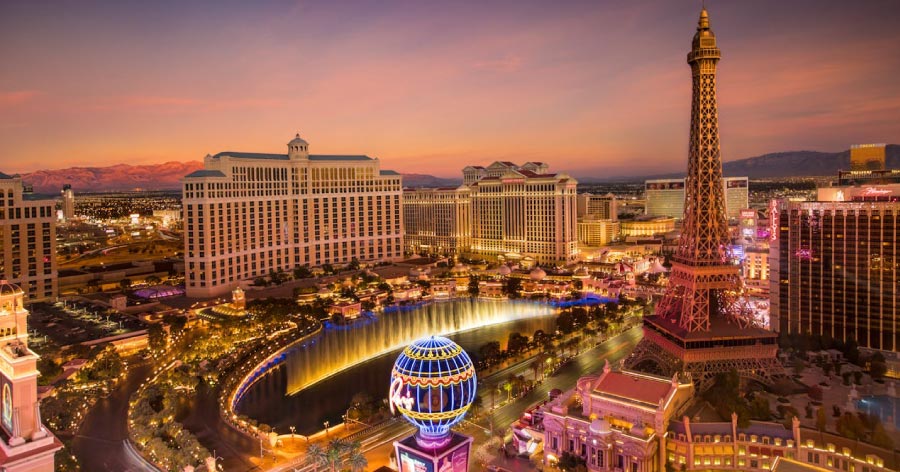 Aerial view of the Las Vegas Strip skyline at sunset featuring the illuminated Bellagio fountains, Eiffel Tower replica, and mountains in the distance.