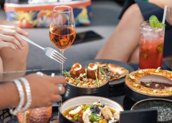 Close-up view of a restaurant table with various upscale dishes and cocktails as friends reach for dishes with their utensils.