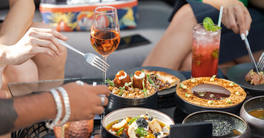 Close-up view of a restaurant table with various upscale dishes and cocktails as friends reach for dishes with their utensils.