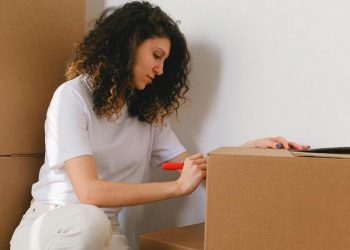 A woman is labeling a cardboard box as she prepares to move out of her college dorm.