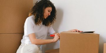 A woman is labeling a cardboard box as she prepares to move out of her college dorm.
