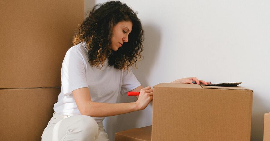 A woman is labeling a cardboard box as she prepares to move out of her college dorm.