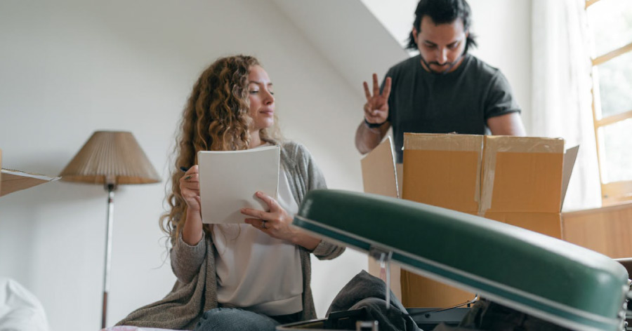 A woman is holding a checklist and looking at her husband, who is holding up three fingers to indicate how many of one item they have already packed in a moving box.