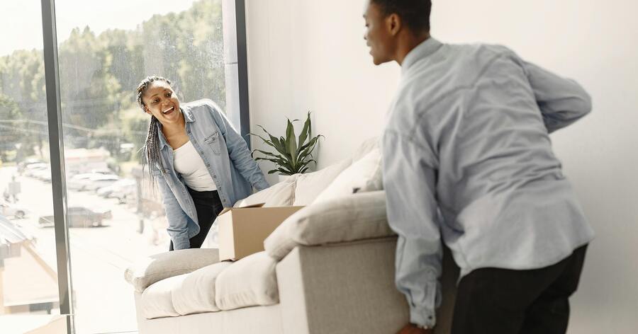A happy couple is moving a couch into their new apartment, which features large windows and lots of natural light.