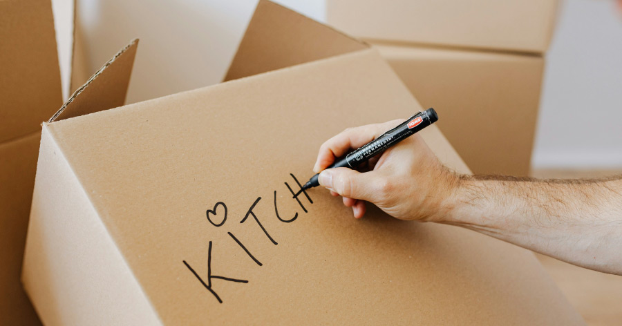 Close-up of a pair of hands writing “KITCHEN” on a moving box with a heart used to dot the “i.”
