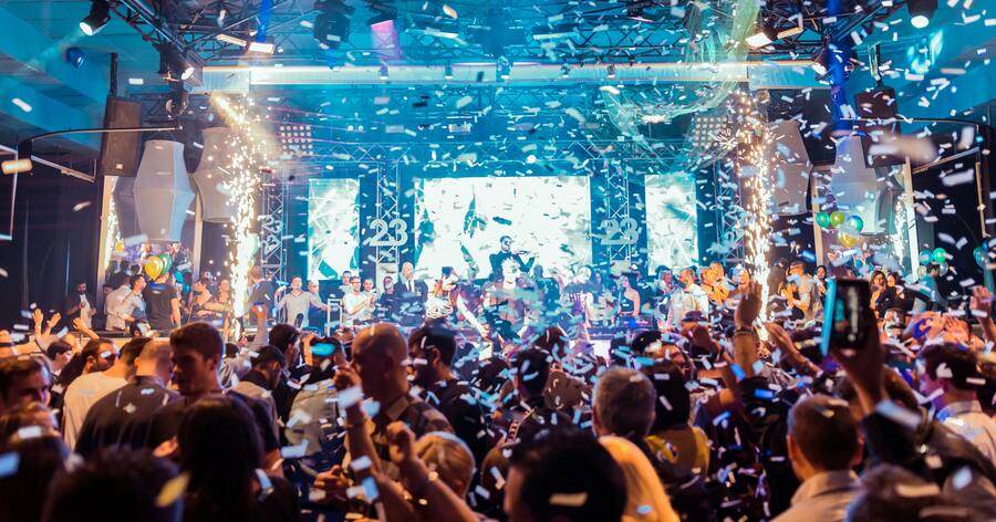A crowd is celebrating at a high-energy nightclub as confetti floats down from the ceiling and stage lights illuminate the space.