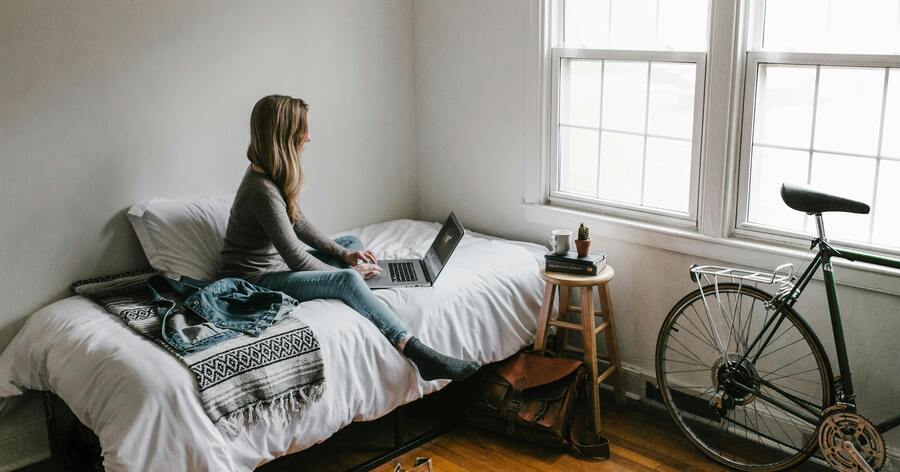 A college student is sitting on the bed in her dorm room, using her laptop to research dorm policies before moving out.