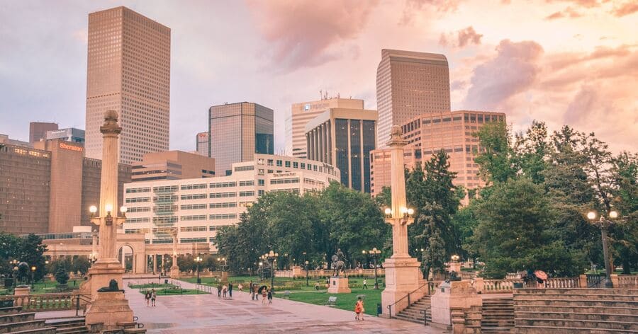 The Denver skyline and Civic Center Park at sunset, with historic monuments, green spaces, and downtown skyscrapers under colorful evening clouds.