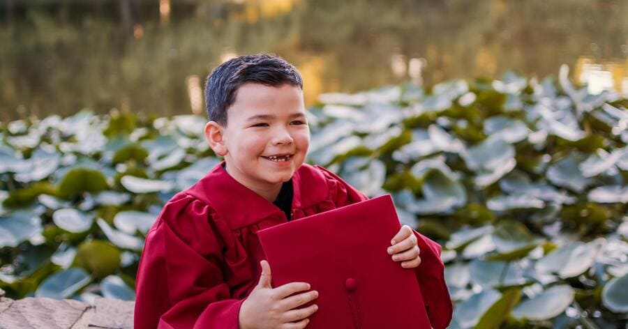 A happy young child in a red graduation gown is holding his graduation cap while standing beside a pond filled with lily pads.