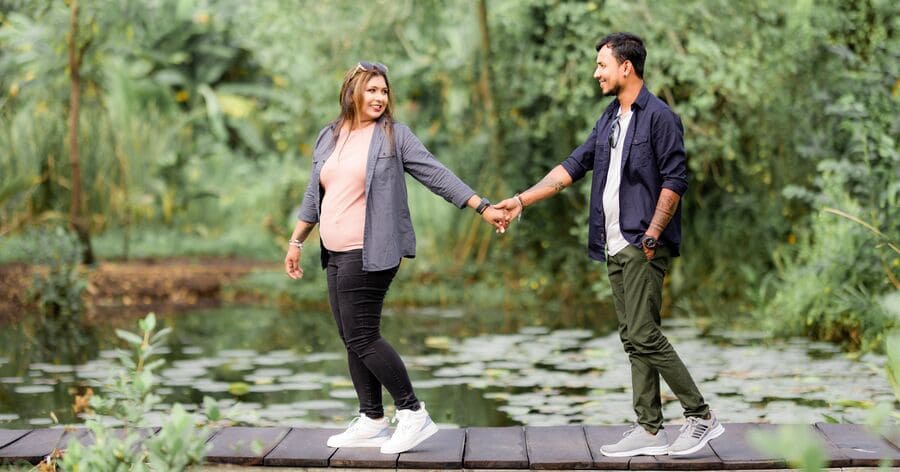 A couple is holding hands while walking across a wooden bridge surrounded by lush greenery and a peaceful pond.