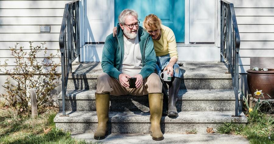 An older couple is sitting together on the front steps of their home, enjoying coffee and conversation.