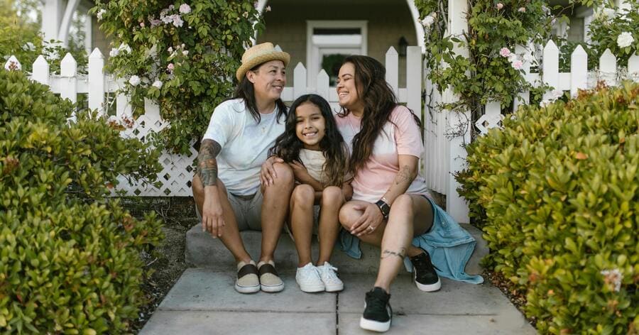 A family of three sitting outside a home with a white picket fence and flowering vines in one of the best Denver neighborhoods for families.