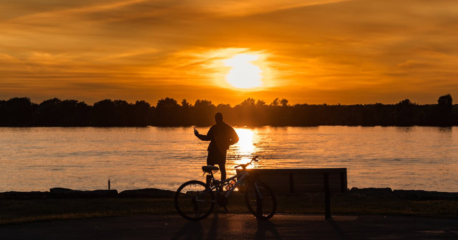 Silhouette of a man standing beside his bicycle looking out on a lake as the sun sets in the distance.