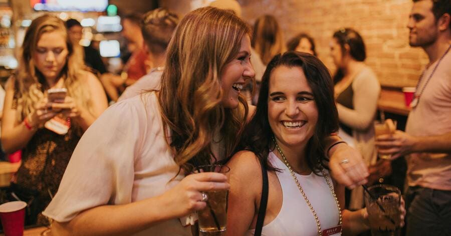Two women laughing and holding drinks while socializing with friends at a crowded bar in one of the best neighborhoods in Denver.