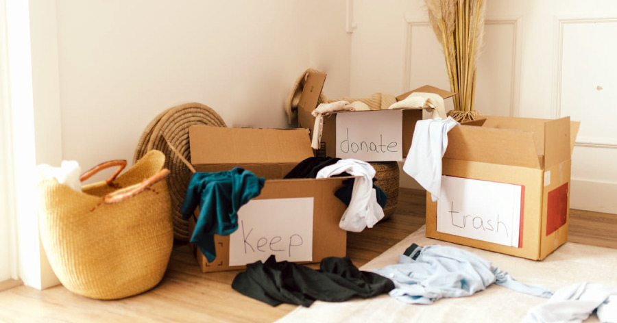 Three cardboard boxes labeled “keep,” “donate,” and “trash” with items of clothing hanging over the edges and a few pieces on the floor.