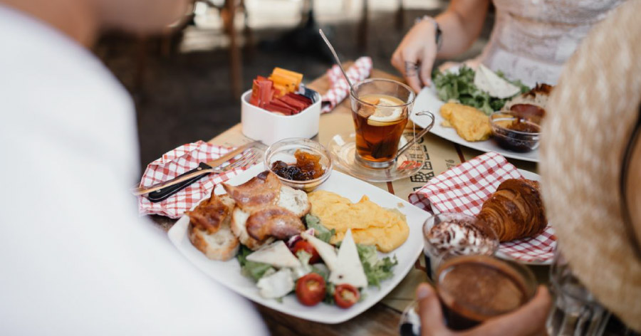 View over a man’s shoulder of the woman sitting across from him and their table, decked out with various dishes, like those found at the best restaurants in Boston.
