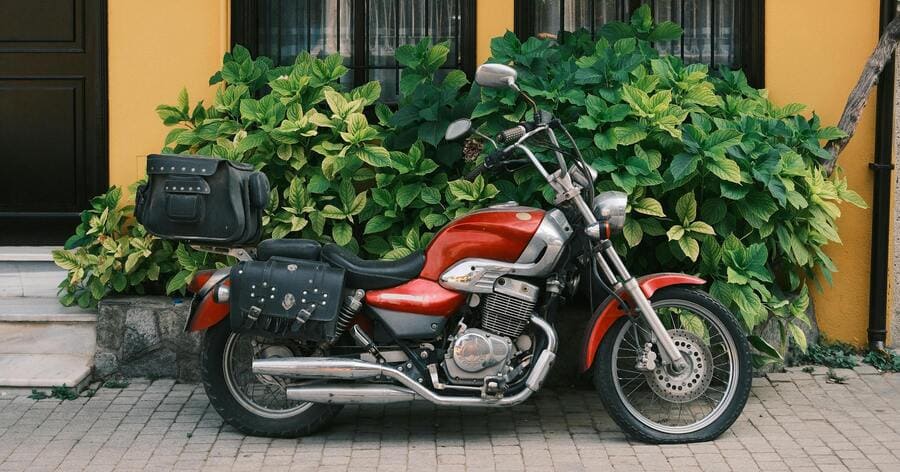 A red cruiser motorcycle parked in front of a yellow house with black trim and lush greenery in a quiet residential neighborhood.