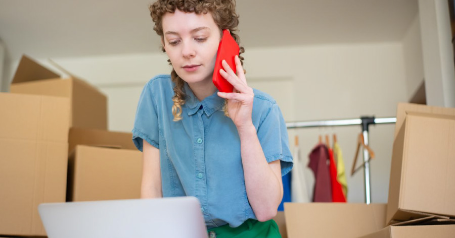 A woman is leaning over her laptop while talking on her cellphone, surrounded by cardboard boxes because she hasn’t found the right small business storage solution.