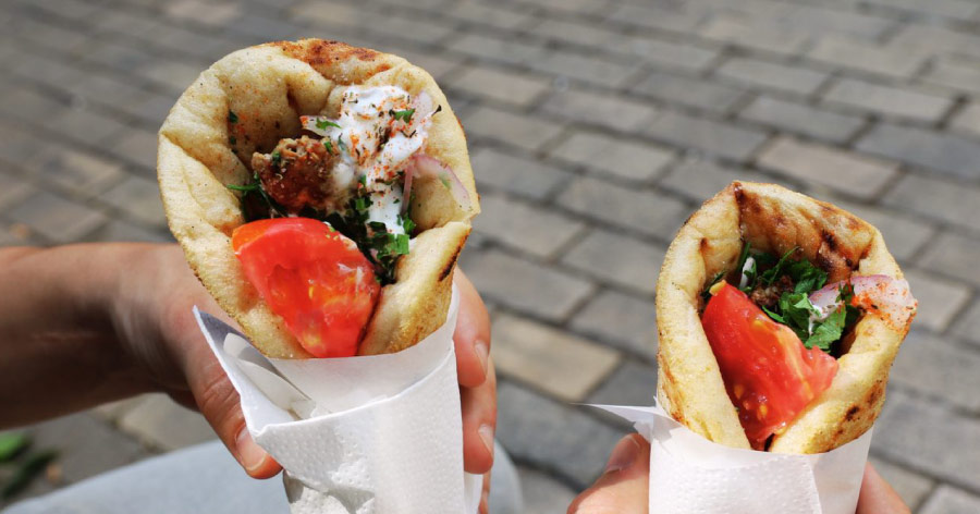 Close-up of hands holding two gyro sandwiches, wrapped in white paper with a brick road in the background.