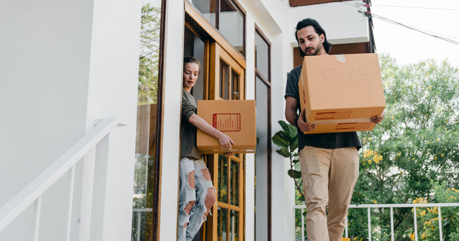 Two people carry moving boxes out of their home after labeling them and clearing a path; simple tips for moving in the summer.